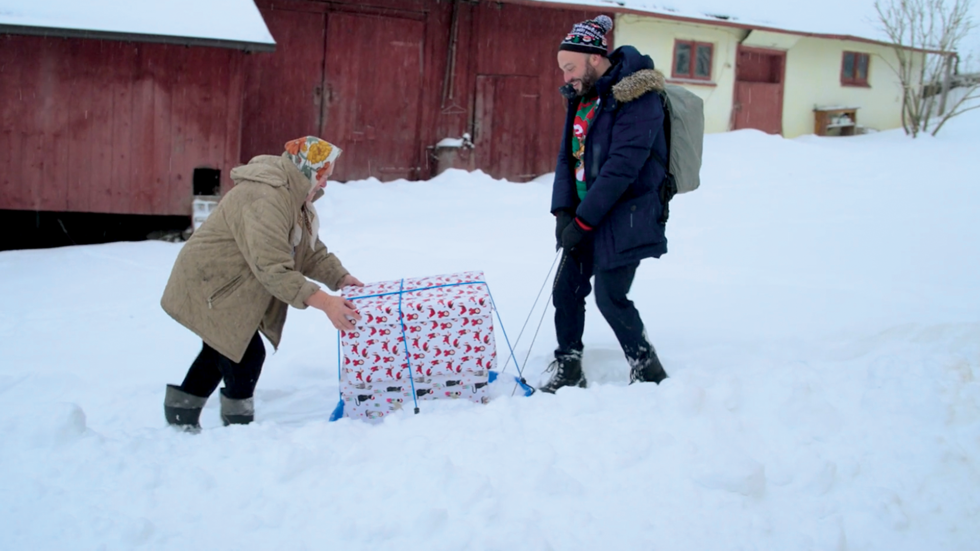 A older woman pushes a wrapped box on a sled through the snow while a middle aged man in a coat pulls the sled